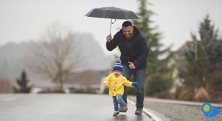 Dad walks his young son in the rain. Their spirits are cheerful and one of them is starting to run away...and Dad is chasing him with a big smile while still holding their umbrella.