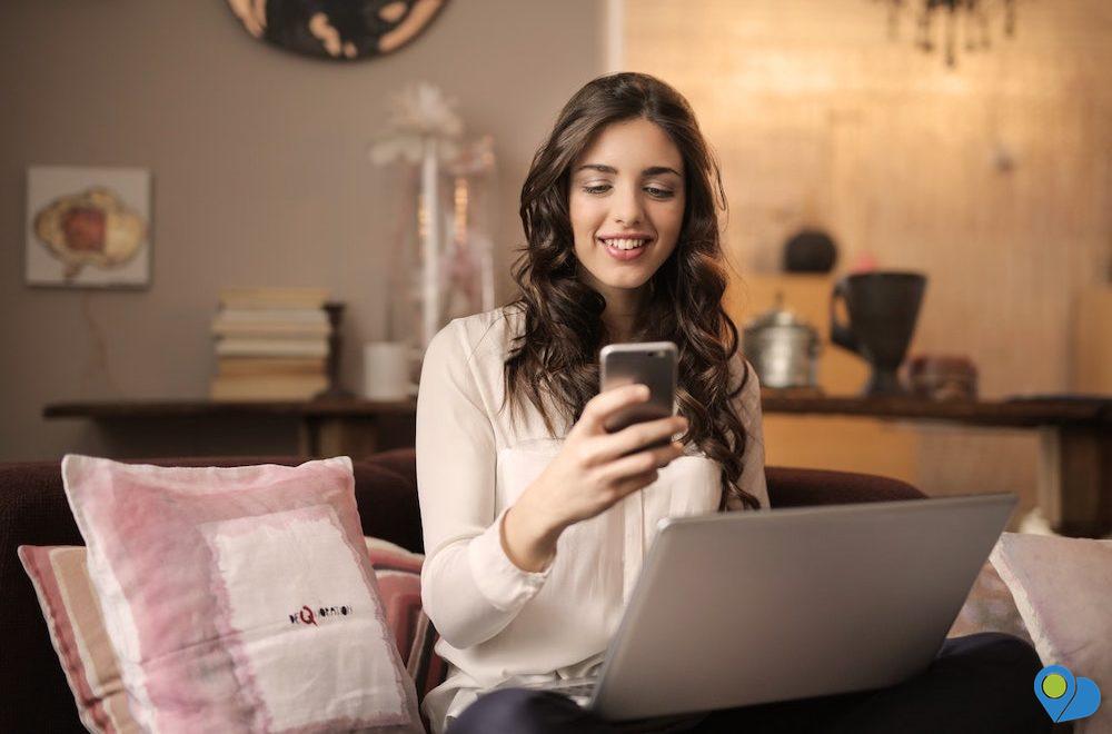 Girl sitting on sofa messaging or checking list on cell phone and laptop