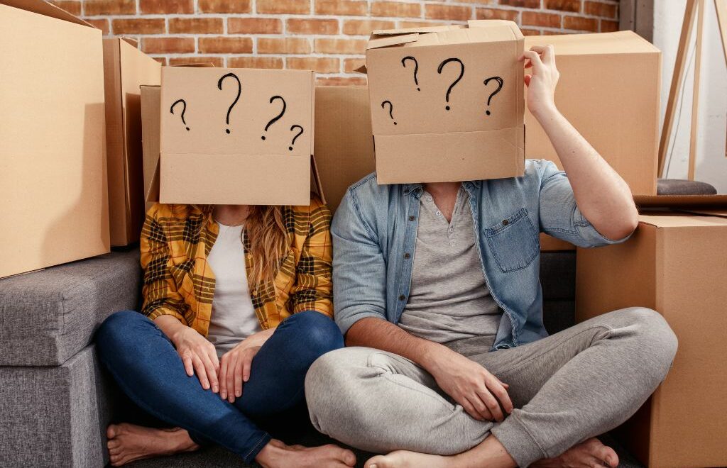 couple sitting on the floor with packing boxes around them and boxes with question marks written on them covering their heads