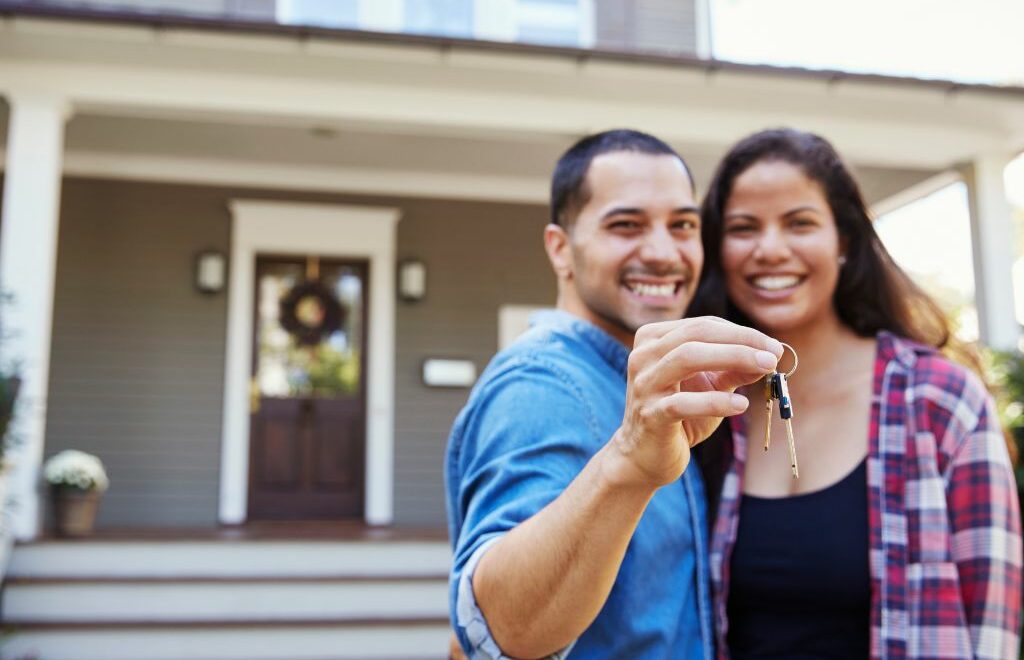 Young couple standing in front of a house holding out keys in front of them