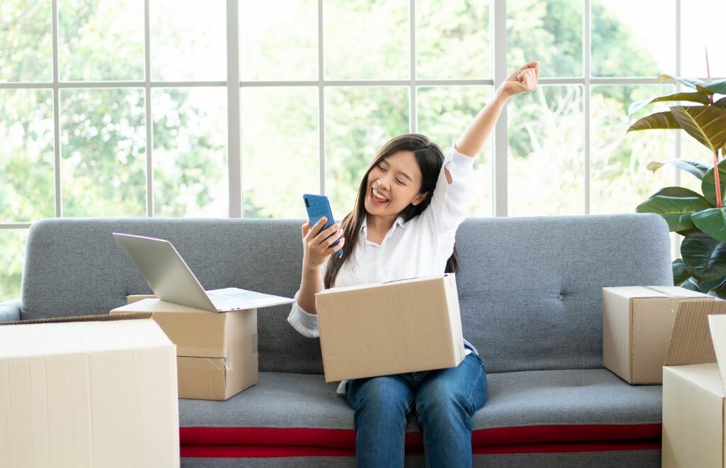 female on sofa with moving boxes, mobile phone and computer celebrating with her arm up in the air