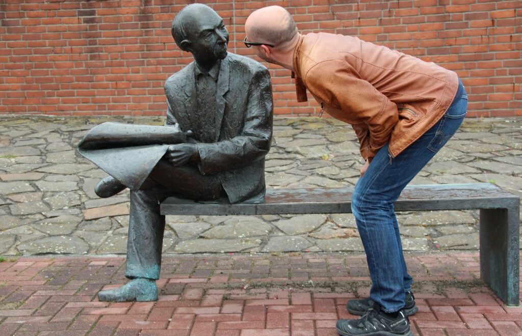 man bending over staring at a sculpture of man sitting on a bench with a newspaper on his lap looking in the direction of the man