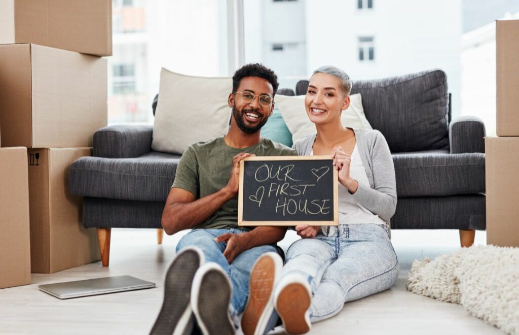 Young couple sitting on the floor between stacks of moving boxes and holding up a little chalkboard saying "Our First House"