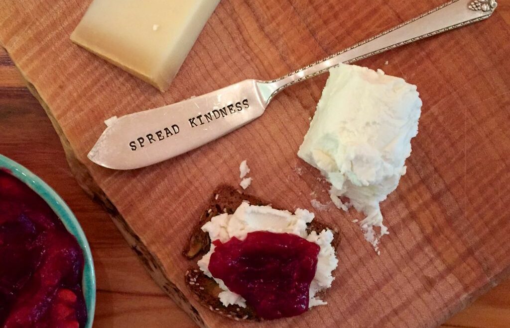 butter knife engraved with "Spread Kindness" with some cheese and toppings around cutting board