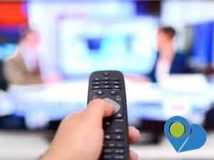 person's finger on a remote control in front of a tv showing a newscast in the background