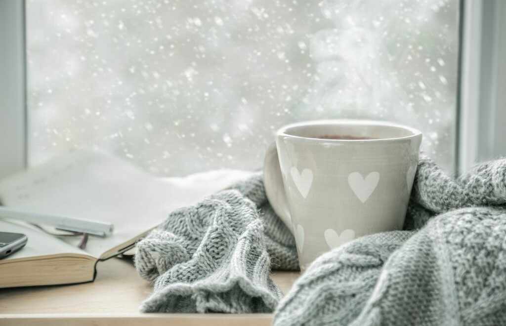 coffee cup nestled in a gray blanket inside a window with snow falling outside