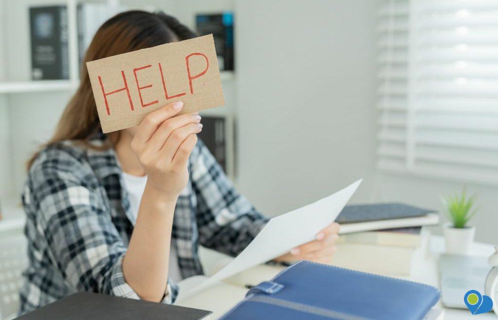 woman sitting at desk holding up "help" sign in front of her face