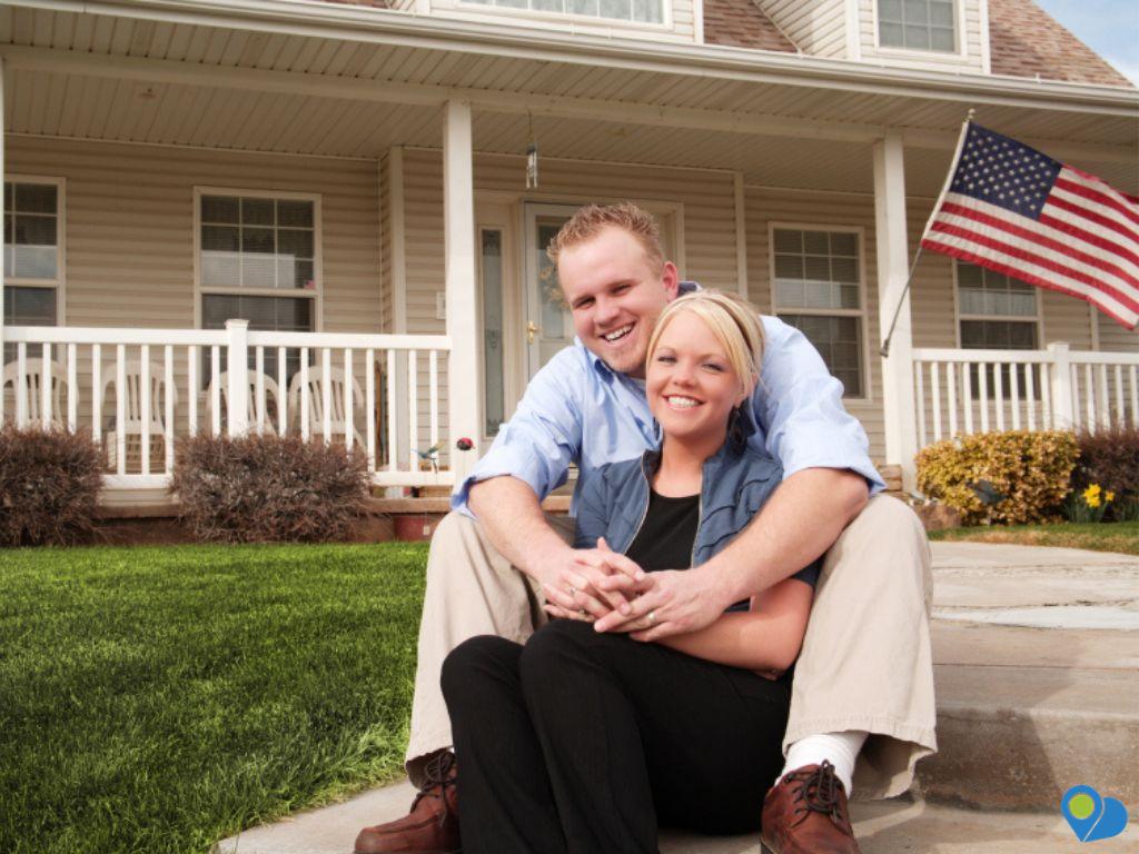 young couple spooning on the front sidewalk of a house with an American flag on the porch